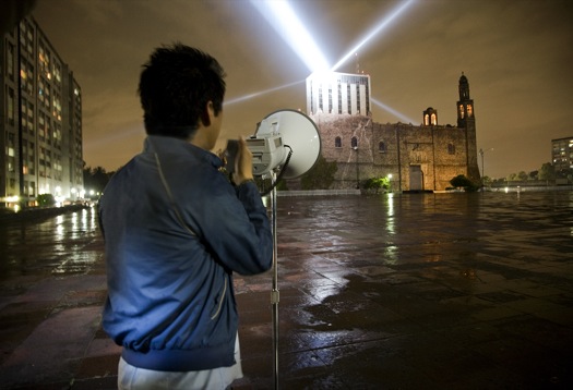 Persona de espaldas que sostiene un megáfono, de fondo el ministerio de Relaciones exteriores de México  y sobre su techo tres haces de luces. 